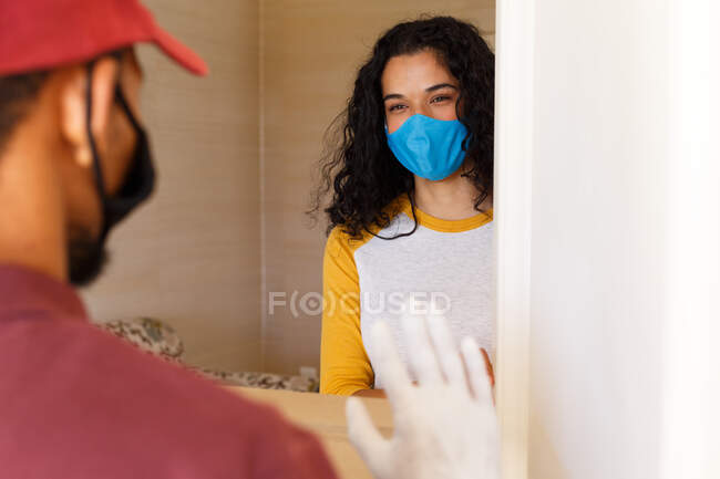 Mujer de raza mixta con una máscara facial saludando al repartidor en la puerta. autoaislamiento en el hogar durante la pandemia de coronavirus covid 19. - foto de stock