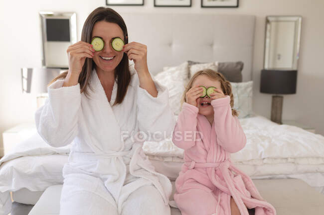 Donna e figlia caucasica si divertono in camera da letto. mettendo fette di cetriolo sugli occhi. godendo di tempo di qualità a casa durante coronavirus covid 19 isolamento pandemico. — Foto stock