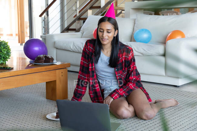 Mixed race woman sitting on floor wearing party hat and using laptop at home. self isolation during covid 19 coronavirus pandemic. — Stock Photo