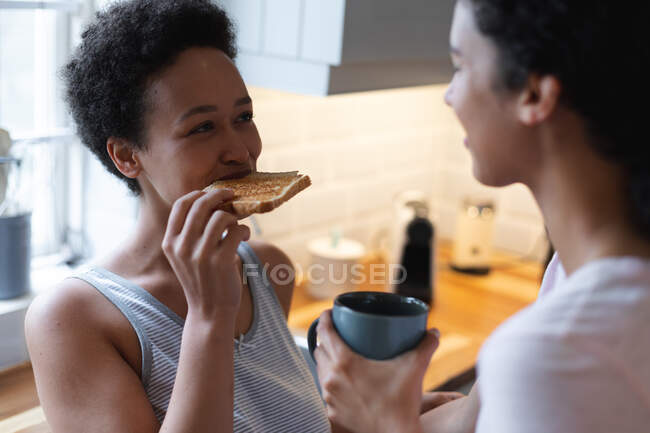 Happy mixed race lesbian couple having toast and coffee for breakfast in kitchen. self isolation quality time at home together during coronavirus covid 19 pandemic. — Stock Photo