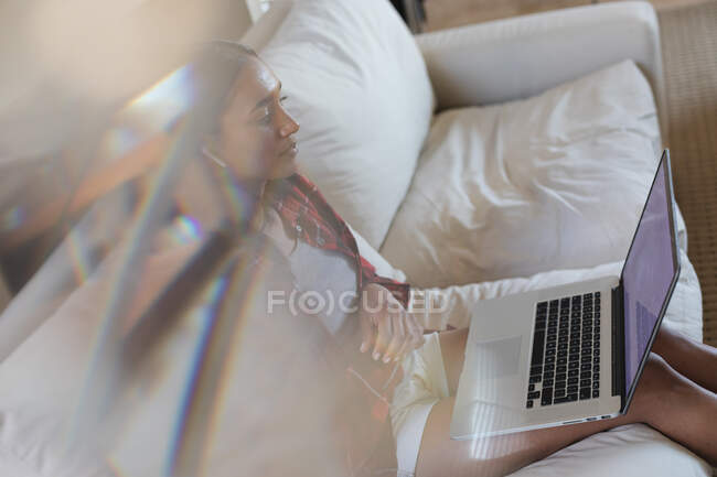 Mixed race woman with wireless earphones on using laptop on couch at home. self isolation during covid 19 coronavirus pandemic. — Stock Photo