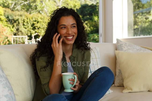 Змішана расова жінка розмовляє на смартфоні, сидячи на дивані у вітальні. самоізоляція в домашніх умовах під час пандемії коронавірусу 19 . — стокове фото