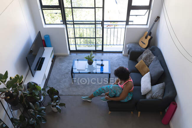 African american woman sitting on couch putting sports shoes on. self isolation at home during coronavirus covid 19 pandemic. — Stock Photo