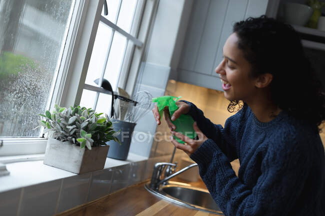 Mixed race woman watering plants in kitchen. self isolation quality family time at home together during coronavirus covid 19 pandemic. — Stock Photo