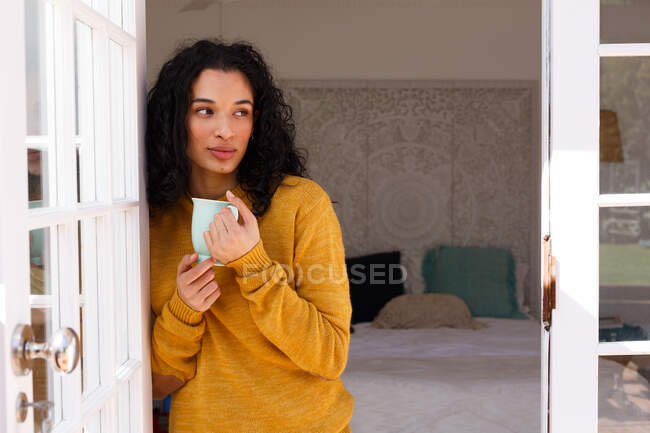 Mixed race woman standing in doors drinking cup of coffee. self isolation during covid 19 coronavirus pandemic — Stock Photo