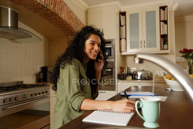 Mujer de raza mixta hablando por teléfono y escribiendo en la cocina. autoaislamiento en el hogar durante la pandemia de coronavirus covid 19. - foto de stock