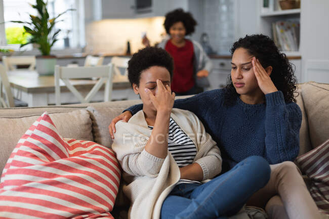 Verärgerte Lesben gemischter Rasse sitzen auf der Couch. Tochter im Hintergrund. Selbst-Isolation Qualität Familienzeit zu Hause zusammen während Coronavirus covid 19 Pandemie. — Stockfoto