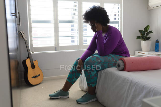 Femme afro-américaine assise sur son lit parlant sur son smartphone. auto-isolement technologie communication à la maison pendant coronavirus covid 19 pandémie. — Photo de stock