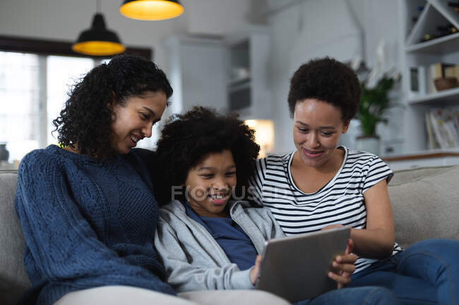 Mixed race lesbian couple and daughter sitting on couch using digital tablet. self isolation quality family time at home together during coronavirus covid 19 pandemic. — Stock Photo