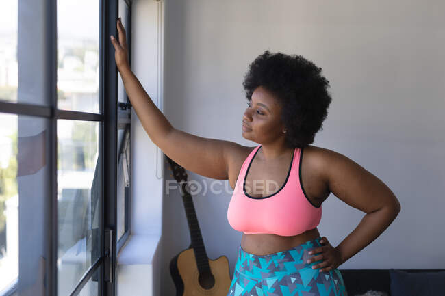 Африканка - американка, одягнена в спортивний одяг, дивиться у вікно. Самоізоляція вдома під час коронавірусу covid 19 пандемії. — стокове фото