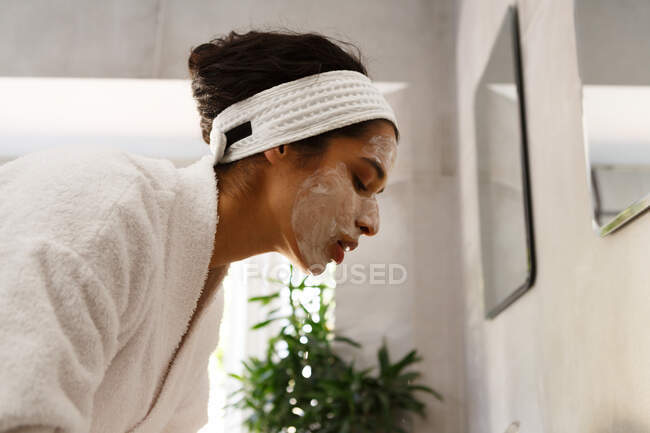 Donna di razza mista che indossa crema per il viso e accappatoio in bagno. auto isolamento a casa durante covid 19 coronavirus pandemia. — Foto stock