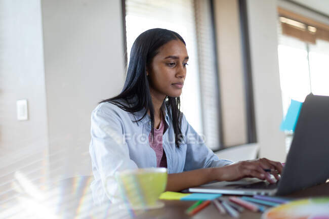 Змішана расова жінка сидить за столом, використовуючи ноутбук, що працює вдома. самоізоляція під час пандемії коронавірусу 19 . — стокове фото