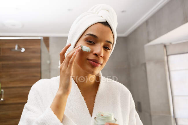 Portrait of mixed race woman applying face cream in bathroom. self isolation at home during covid 19 coronavirus pandemic. — Stock Photo