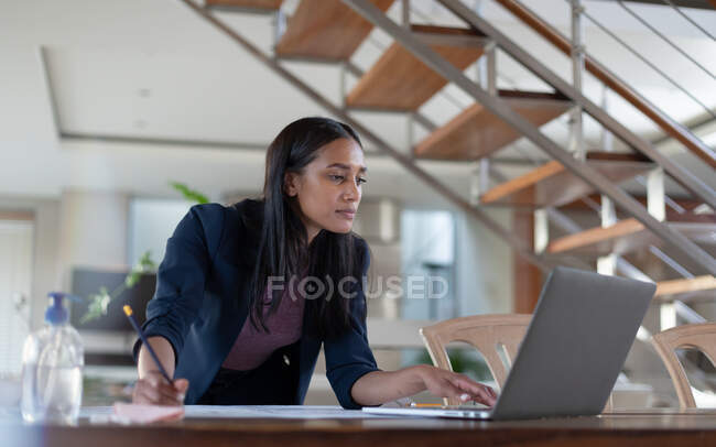 Mujer de raza mixta sentada en la mesa usando un portátil, escribiendo, trabajando en casa. autoaislamiento durante la pandemia de coronavirus covid 19. - foto de stock