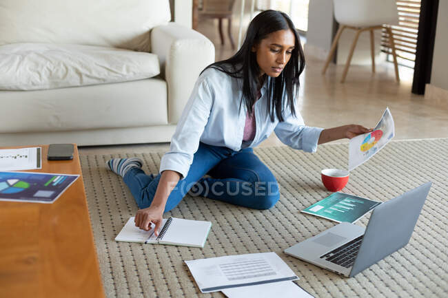 Gemischte Rassenfrau, die mit Laptop auf dem Boden sitzt und Dokumente in der Hand hält, die zu Hause arbeiten. Selbstisolation während der Covid 19 Coronavirus-Pandemie. — Stockfoto