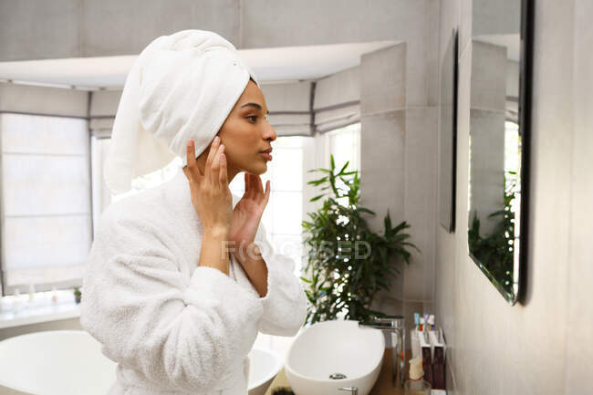 Змішана расова жінка, дивлячись у дзеркало, наносить крем для обличчя у ванній кімнаті. самоізоляція в домашніх умовах під час пандемії коронавірусу 19 . — стокове фото