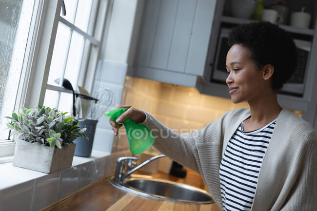 Змішана раса жінка поливає рослини на кухні. самоізоляція якість сімейного часу вдома разом під час пандемії коронавірусу 19 . — стокове фото