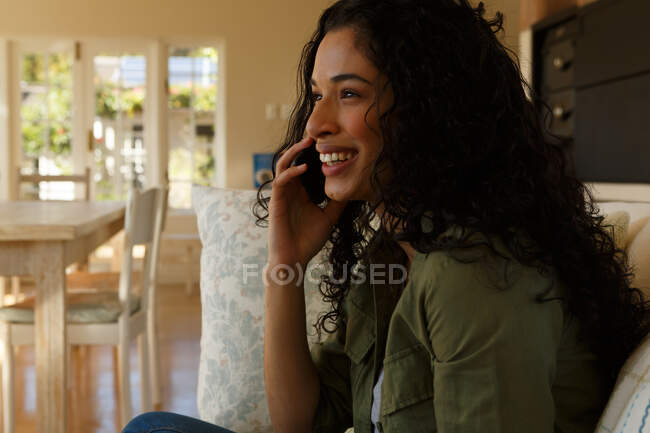Mulher de raça mista falando no smartphone sentado no sofá na sala de estar. auto-isolamento em casa durante a pandemia do coronavírus covid 19. — Fotografia de Stock