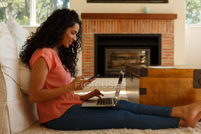 Mujer de raza mixta con teléfono inteligente y portátil sentado en el suelo en la sala de estar. autoaislamiento en el hogar durante la pandemia de coronavirus covid 19. - foto de stock