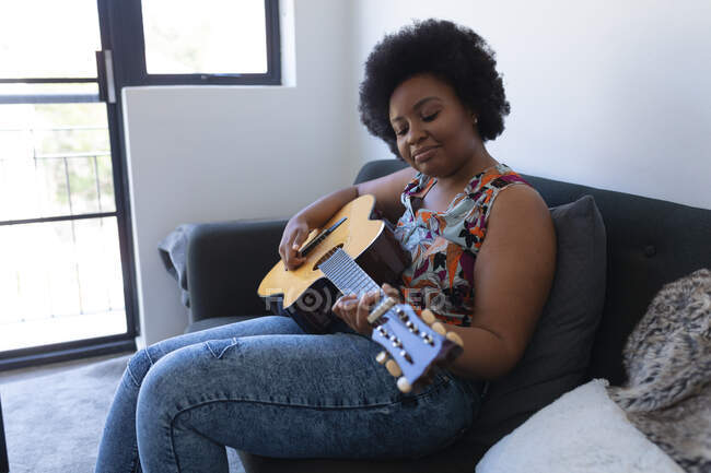 Усміхнена афро-американська жінка, яка сидить на дивані, грає на акустичній гітарі. Музика для самоізоляції під час коронавірусу covid 19 пандемії. — стокове фото