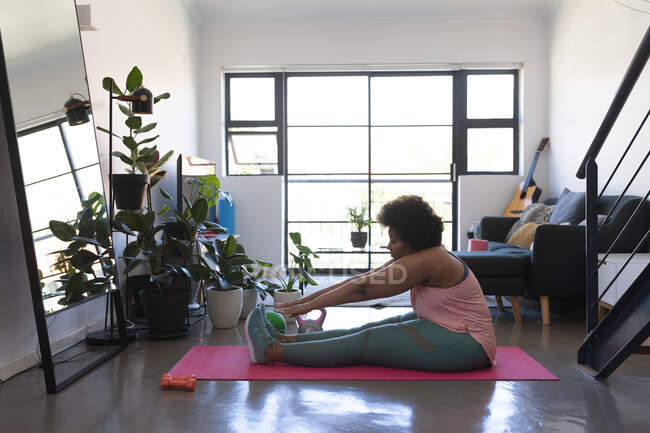 Mujer afroamericana sentada en una alfombra de ejercicio haciendo ejercicio. autoaislamiento fitness en casa durante coronavirus covid 19 pandemia. - foto de stock