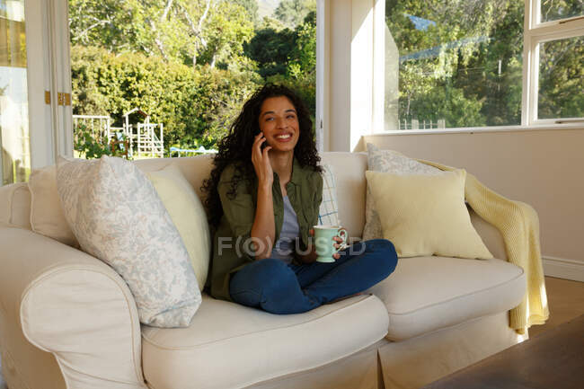 Mujer de raza mixta hablando en el teléfono inteligente sentado en el sofá en la sala de estar. autoaislamiento en el hogar durante la pandemia de coronavirus covid 19. - foto de stock