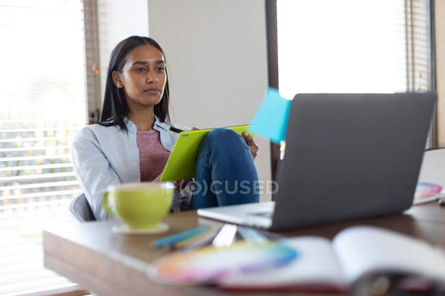 Mulher de raça mista sentada à mesa segurando prancheta trabalhando em casa. auto-isolamento durante a pandemia do coronavírus covid 19. — Fotografia de Stock