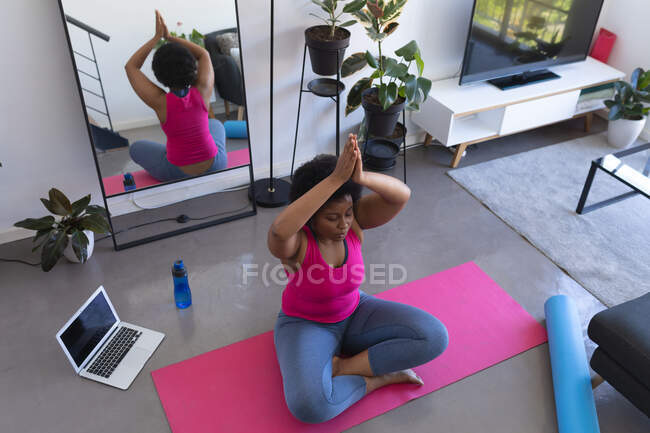 Африканська жінка, яка займається медитацією йоги, сидить на маті в спортивному одязі. Ноутбук на задньому плані. Технологія самоізоляції здорового самоізоляції вдома під час коронавірусу covid 19 пандемії. — стокове фото