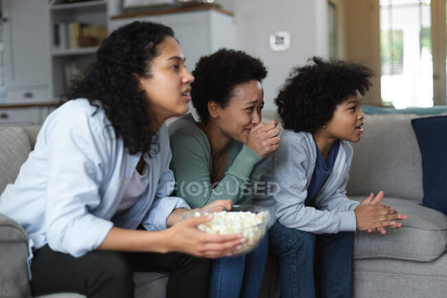 Смешанная раса лесбийская пара и дочь сидят на диване, смотрят телевизор и едят попкорн. самоизоляция качество семейное время дома вместе во время коронавируса ковид 19 пандемии. — стоковое фото