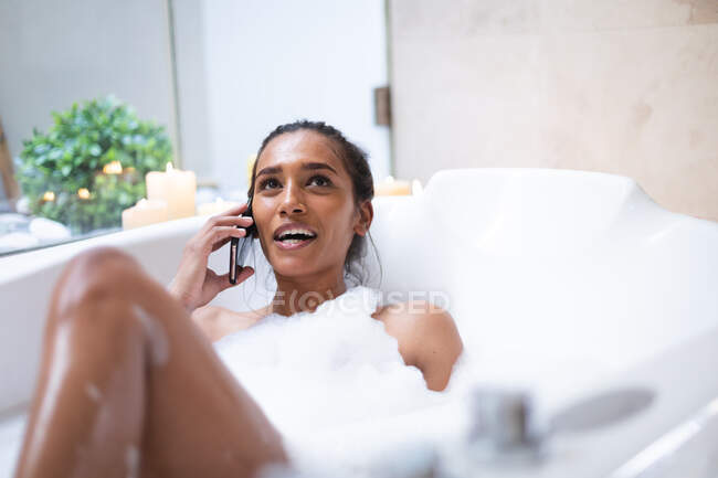 Mujer de raza mixta tumbada en el baño relajante y el uso de smartphone. autoaislamiento durante la pandemia de coronavirus covid 19. - foto de stock