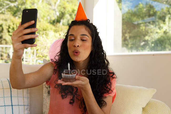 Mujer de raza mixta celebrando cumpleaños teniendo chat de vídeo en el teléfono inteligente. Usando sombrero de fiesta y soplando una vela en un panecillo. autoaislamiento en el hogar durante la pandemia de coronavirus covid 19. - foto de stock