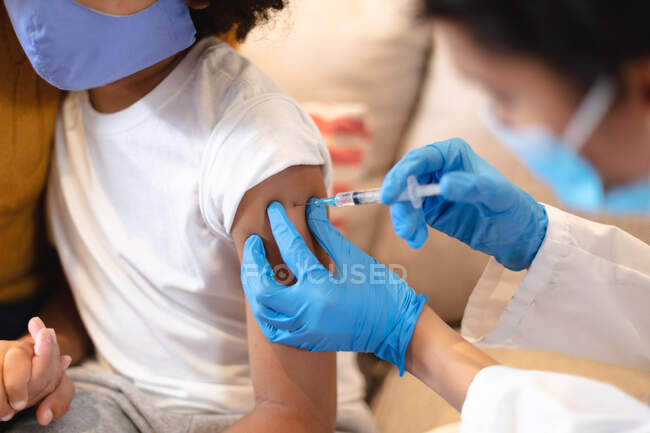 Chica de raza mixta con máscara facial que recibe la vacuna contra la gripe por parte de una doctora. autoaislamiento en casa durante la pandemia del coronavirus covid 19 - foto de stock