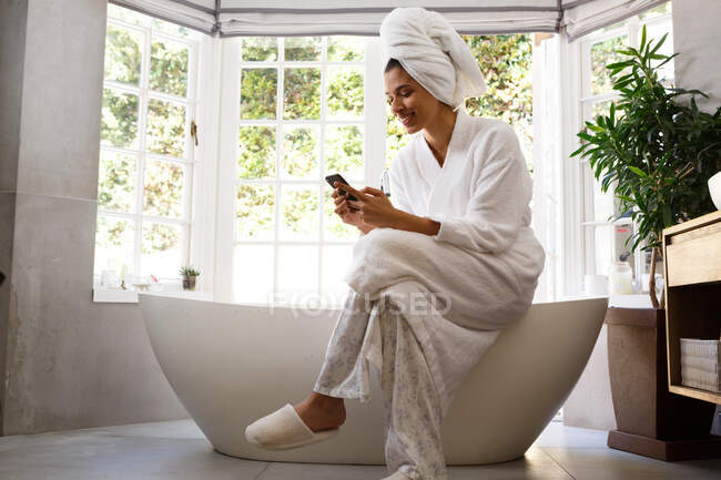 Mixed race woman wearing bathrobe sitting on bathtub using smartphone. self isolation at home during covid 19 coronavirus pandemic. — Stock Photo