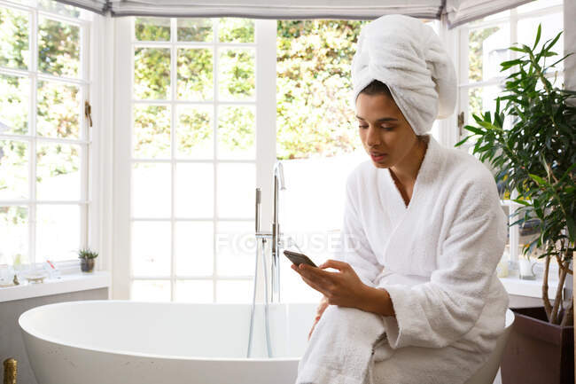 Femme de race mixte portant un peignoir assis sur la baignoire à l'aide d'un smartphone. auto-isolement à la maison pendant la pandémie de coronavirus covid 19. — Photo de stock