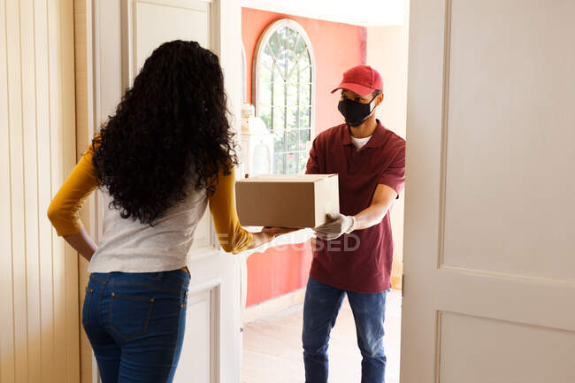 Mixed race woman wearing face mask receiving a package. self isolation during covid 19 coronavirus pandemic — Stock Photo