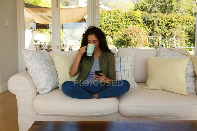 Смешанная расовая женщина пьет кофе, используя смартфон, сидя на диване в гостиной. самоизоляция в домашних условиях во время пандемии 19 коронавируса. — стоковое фото