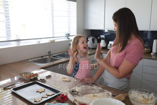 Madre e hija caucásicas divirtiéndose horneando en la cocina, chocando los cinco. disfrutando de tiempo de calidad en casa durante coronavirus covid 19 pandemia de bloqueo. - foto de stock