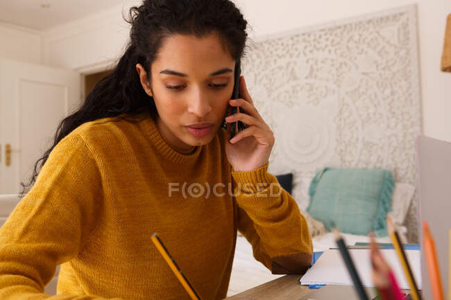 Mujer de raza mixta hablando en el teléfono inteligente sentado en el escritorio de escritura. autoaislamiento en el hogar durante la pandemia de coronavirus covid 19. - foto de stock