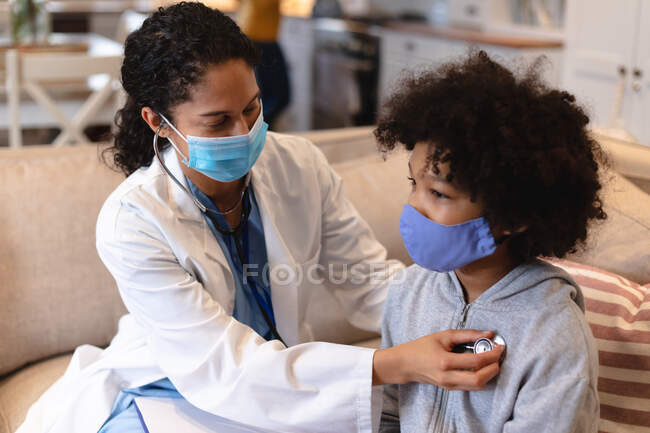 Chica de raza mixta que usa mascarilla siendo examinada por una doctora de raza mixta sentada en un sofá. aislamiento en casa durante la pandemia de coronavirus covid 19. - foto de stock