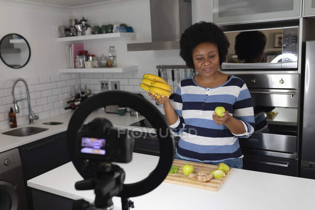 Vloggeuse afro-américaine enregistrant une vidéo dans la cuisine. couper des légumes. auto-isolement technologie communication à la maison pendant coronavirus covid 19 pandémie. — Photo de stock