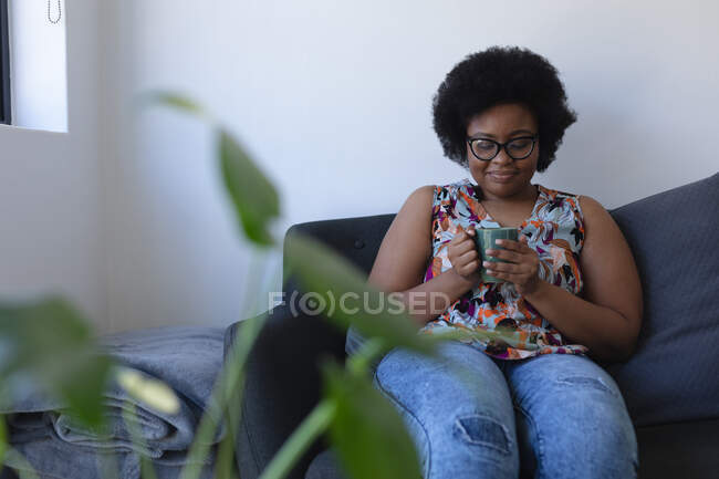 Sorrindo afro-americana sentada no sofá desfrutando de uma xícara de café. auto-isolamento em casa durante coronavírus covid 19 pandemia. — Fotografia de Stock