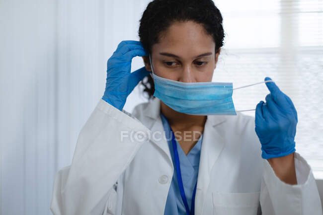 Портрет жіночого лікаря змішаної раси, який дивиться на камеру і одягає маску обличчя. Якість самоізоляції вдома під час коронавірусу covid 19 пандемії. — стокове фото
