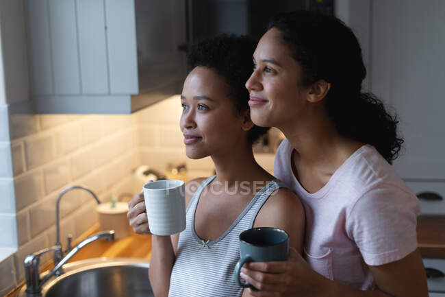 Sorrindo casal lésbico de raça mista bebendo café e abraçando na cozinha. auto isolamento tempo de qualidade em casa juntos durante coronavírus covid 19 pandemia. — Fotografia de Stock