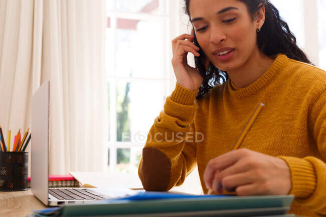 Mixed race woman talking on smartphone writing sitting at desk with laptop. self isolation at home during covid 19 coronavirus pandemic. — Stock Photo