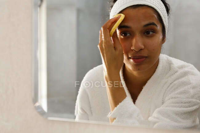 Mixed race woman reflected in mirror cleansing face in bathroom. self isolation at home during covid 19 coronavirus pandemic. — Stock Photo