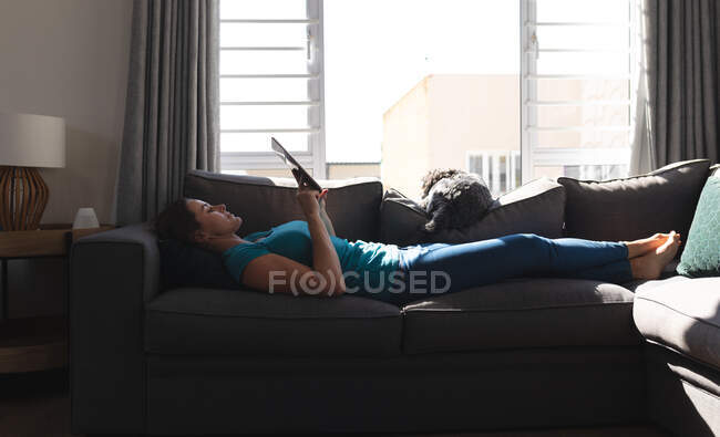Улыбающаяся кавказка лежит на диване в гостиной с помощью планшета. наслаждаясь временем дома во время коронавирусного ковида 19 пандемического блокирования. — стоковое фото