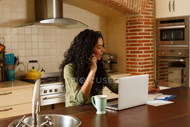 Mujer de raza mixta hablando por teléfono usando el ordenador portátil en la cocina. autoaislamiento en el hogar durante la pandemia de coronavirus covid 19. - foto de stock