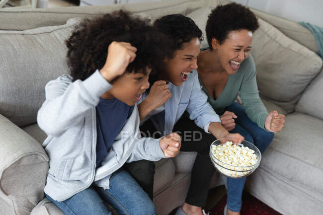 Mixed race lesbian couple and daughter sitting on couch watching tv and eating popcorn. cheering together. self isolation quality family time at home together during coronavirus covid 19 pandemic. — Stock Photo