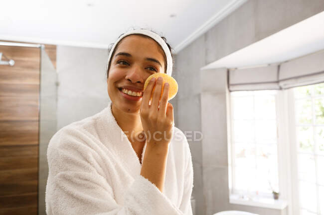 Mixed race woman looking in mirror cleansing face in bathroom. self isolation at home during covid 19 coronavirus pandemic. — Stock Photo