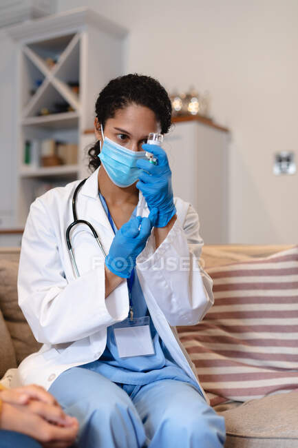 Médica de raça mista usando máscara facial sentada no sofá. enchendo uma seringa. auto isolamento tempo de qualidade em casa juntos durante coronavírus covid 19 pandemia. — Fotografia de Stock